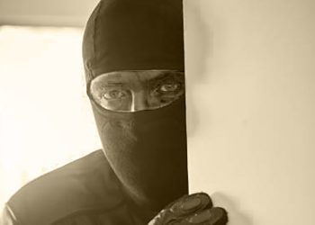 A sinister man wearing all black and made unrecognizable by a balaclava, peeps round a door after breaking into a home.