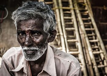 Portrait of Indian elder man with traditional bindi as a third eye, white beard and bamboo ladders on the background in Mysore, Karnataka, India. Concerned expression