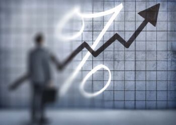 A businessman, standing while holding a briefcase, looks up at a chart on a wall that indicates interest rates are rising.